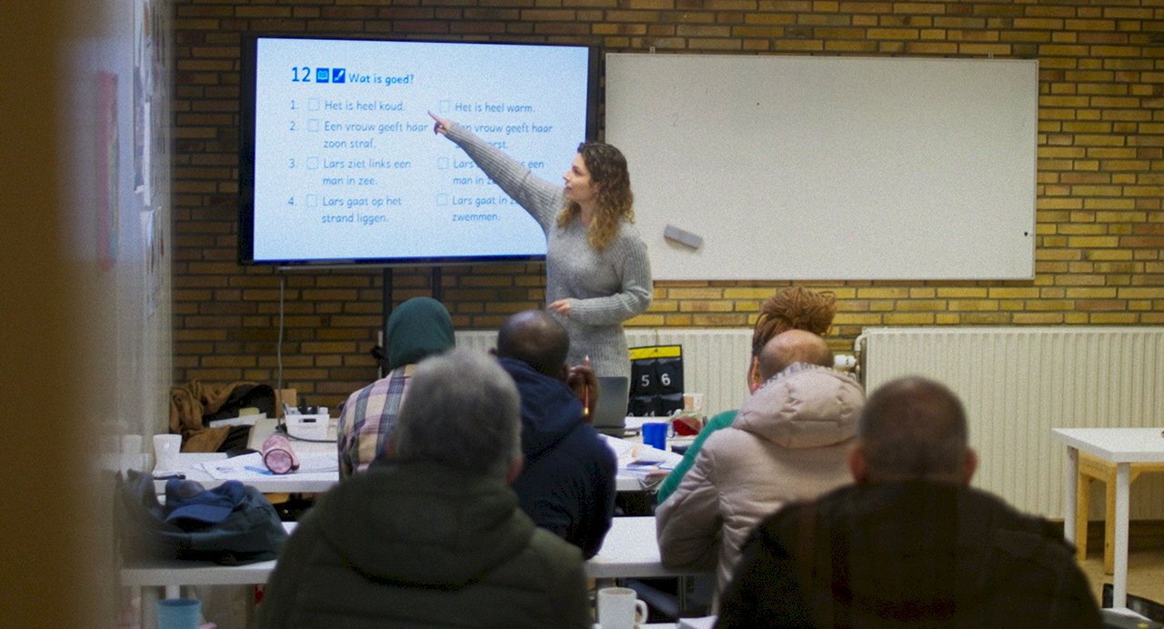 Docent geeft taalles en wijst naar het scherm terwijl cursisten meekijken in het klaslokaal